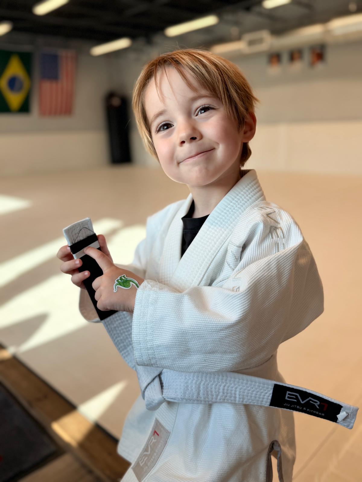 A young student in a white gi smiling while holding a rolled-up belt on the academy mats