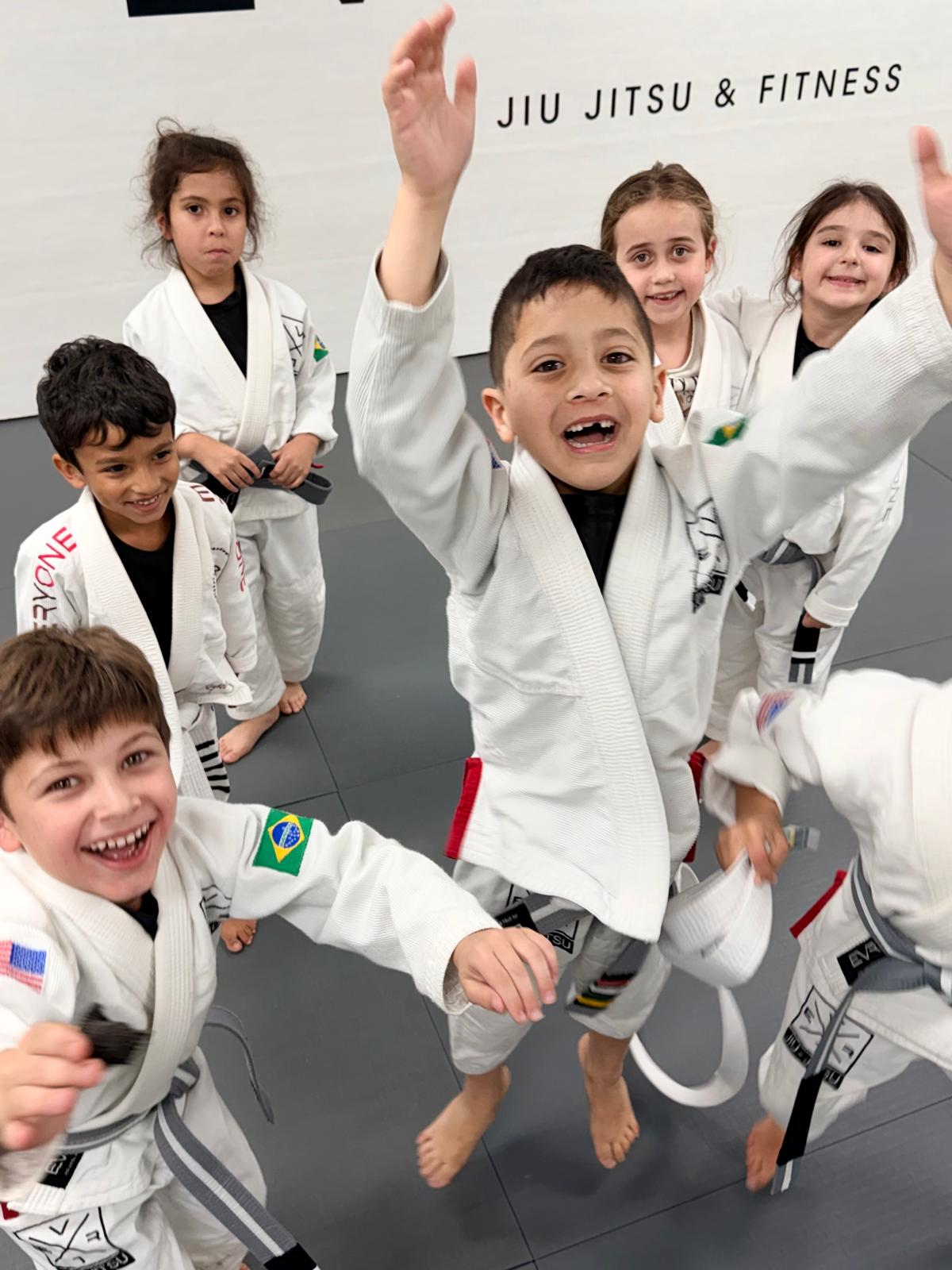 Kids in white gis smiling and reaching up on the mats at Everyone Jiu Jitsu